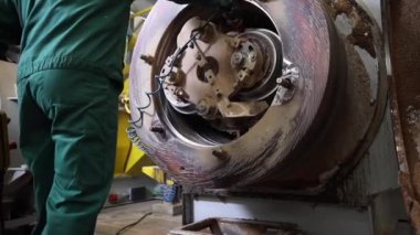 Bir fabrika işçisi büyük dairesel bir makine bileşenini inceliyor. Ayarlar arasında aşınmış makineler, sarı bir yapı ve beton zemin yer alıyor..