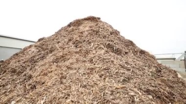 Önplanda, endüstriyel yapılara ve arka planda bulutlu gökyüzüne karşı duran görünür desenli büyük bir odun yığını var..