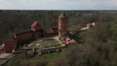 Silindirik bir kulesi, saati ve görünür kalıntıları olan kırmızı tuğlalı kale, bulutlu bir gökyüzünün altında dağınık binaları ve yolları olan bir orman manzarasının ortasına kurulmuştur..