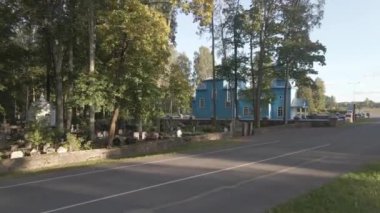 Beyaz pencereli mavi ahşap bir kilise, sonbahar yapraklı uzun ağaçlarla çevrili taş bir duvarla çevrili bir mezarlığın yanında duruyor..