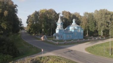 Gümüş çatılı mavi ahşap bir kilise, merkez kubbe, ve çan kulesi kaldırımlı bir yolun yanında duruyor, uzun ağaçlar ve taş bir çitle çevrili..