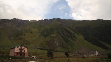 Çimen ve bitki örtüsü olan engebeli dağ yamaçları, dağınık kırsal binalar, yeşil çatılı ev, ağaçlar, park edilmiş araçlar, ve parçalı bulutlu bir gökyüzü..