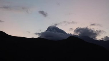 Kazbek Dağı Kafkasya Dağları 'nda belirgin bir şekilde yükselir ve zirvesi kısmen bulutlarla gizlidir. Yuvarlanan tepeler ve loş bir gökyüzü sahneyi kareler..