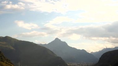 Parçalı bulutlu bir gökyüzünün altında belirgin sivri dağ zirvesi, eğimli tepeler, seyrek bitki örtüsü ve Georgia 'daki üste küçük bir yerleşim yeri..