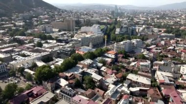Özgürlük Anıtı, ağaç sıralanmış sokaklar, tarihi ve modern mimari ve açık gökyüzünün altındaki tepeler yer alan Tiflis şehir manzarası.