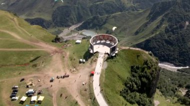 Dağlar, nehir, göl, ziyaretçiler ve yukarıda bir paraglider ile çevrili Gudauri, Gürcistan 'daki bir uçurumda renkli duvar resimleri olan dairesel bir anıt..