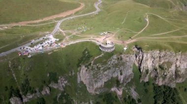Çayırların, patikaların, çadırların ve gökyüzünde paraşütçülerin olduğu dolambaçlı bir yolun bulunduğu bir uçurumun üzerindeki dairesel taş anıt..