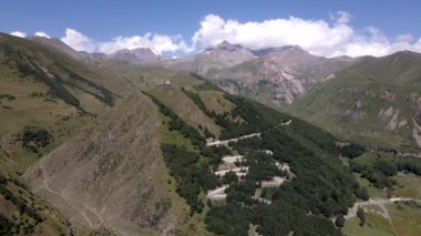 Gürcistan 'ın dağlık arazisinde Gergeti Trinity Kilisesi, dolambaçlı yollar, ormanlık yamaçlar ve bulutlu gökyüzünün altındaki karlı tepeler yer alıyor..