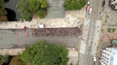 Riga, Letonya 'da düzenlenen bir maratona ait hava perspektifinde, parkın yakınındaki bir yolda katılımcılar, su üzerindeki bir köprü ve yakındaki kentsel yapılar yer alıyor..