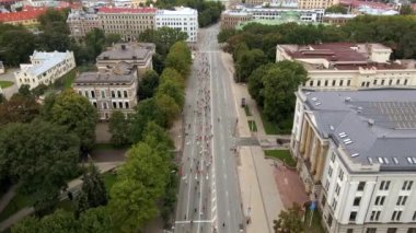 Katılımcılar Letonya 'nın Riga kentinde geniş bir cadde boyunca ilerlerken, kentteki tarihi binalar, ağaçlar ve yeşil alanlar tarafından çevreleniyor..