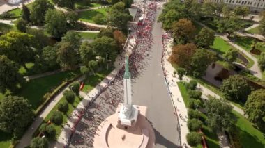Letonya 'nın Riga kentindeki Özgürlük Anıtı' nın hava perspektifi maraton koşucuları, seyirciler, ağaçlar, patikalar ve küçük su hatlarıyla çevrili..