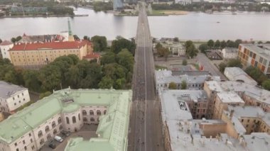 Koşucular Letonya 'nın Riga kentindeki Vansu Köprüsü' nü bir maraton sırasında geçiyorlar. Sahnede Riga Kalesi, tarihi binalar ve Daugava Nehri kıyısındaki modern yüksek binalar yer alıyor..