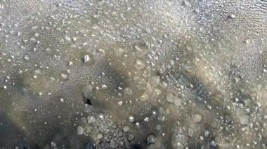 Yarı saydam buzun içinde sıkışmış hava kabarcıkları, düzensiz dokular ve altında koyu bir katman bulunan donmuş bir yüzey hakkında detaylı bir görüş..