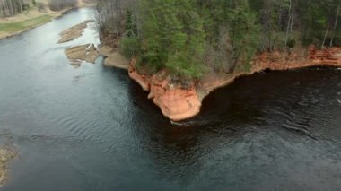 Koyu renkli su kıvrımları olan bir nehir kırmızı kumtaşı bir uçurum etrafında yeşil ağaçlarla kaplı, seyrek bitki örtüsü ve açık nehir kıyılarıyla çelişiyor..
