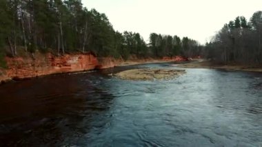 Kırmızımsı kahverengi uçurumlar ve yaprakları dökülen ağaçlar arasında akan kıvrımlı bir nehir. Dalgalanan su ve yakınındaki seyrek bitki örtüsüyle birlikte..