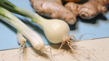 Taze yeşil soğanlar, kökleri olan beyaz bir soğan ve açık bir yüzeyde doğal dokuları vurgulayan, mavi arkaplan üzerine zencefil kökü parçaları..