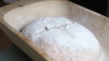 Bir somun çoklu tahıllı ekmek hamuru, un tozuyla kaplı ahşap bir yalıtım tepsisinin içinde duruyor. Çapraz puanlama ve doğal ışıklandırma sanatsal süreci vurguluyor.