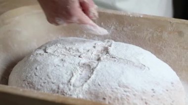 Bir el ahşap bir tepside, un tozuyla kaplanmış ve haçla çizilmiş yuvarlak hamurla çalışır. Bulanık arkaplan ekmek yapma sürecini vurgular.