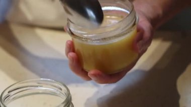 Benekli bir tezgahın üzerindeki temiz cam kavanoz yavaş bir dereye dökülen akışkan, açık renkli bir maddeyle doludur. Yakınlarda renkli bir kapak var..