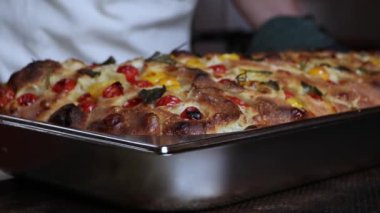 Üzerinde kiraz domatesli, fesleğenli, zeytinyağlı, beyaz önlüklü ve siyah fırın eldivenli biri tarafından bir tepside tutulan altın kahverengi Focaccia ekmeği..