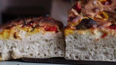 Taze pişmiş Focaccia ekmeği havadar içini göstermek için dilimlenmiş, üzerinde kavrulmuş kiraz domatesleri, sarı biberler ve pişirme tepsisindeki şifalı otlar var..