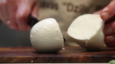 Eller ahşap bir kesme tahtasının üzerinde mozzarella peyniri topunu dilimler. Bıçak yumuşak, parlak yüzeyi kesiyor, bulanık bir arka planı var..