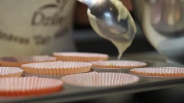 Bir kaşık hamur hamurunu metal bir fırın tepsisiyle pembe kağıt kaplamalarına aktarır. Mutfak arka planında bulanık bir karıştırma kabı ve kabı görülüyor..