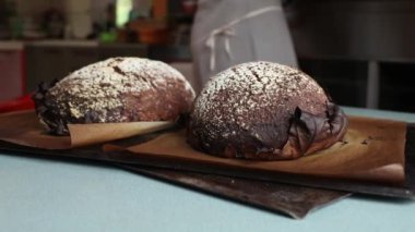 Parşömen astarlı tepsilerde un tozuyla kaplı iki somun siyah ekmek dinleniyor. Bulanık mutfak arka planında tezgah, çatal bıçak ve önlük giyen biri vardır..