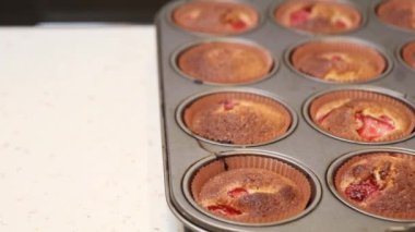 Her birinin üstünde kırmızı meyve parçaları olan metal bir teneke kutuda taze pişmiş muffinler benekli açık renkli bir tezgahın üzerine yerleştirilmiş..
