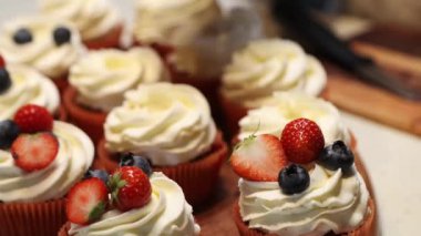 Üzerinde beyaz krema, çilek ve yaban mersiniyle süslenmiş kapkekler kesme tahtası ve bıçakla bir yüzeye dizilmiş..