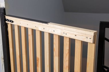 Dikey çıtçıtlı ahşap bir güvenlik kapısı ve merdivenlerin üstüne siyah metal bir menteşe yerleştirilmiş. Yakınlarda gri bir duvar ve siyah metal korkuluklar var..