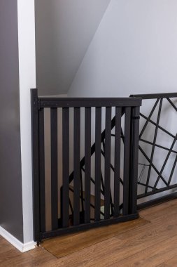 Dikey çıtçıtlı ve mandal mekanizmalı siyah güvenlik kapısı, geometrik metal korkuluklara bitişik. Gri ve beyaz duvarlar ahşap döşemeyi tamamlıyor.