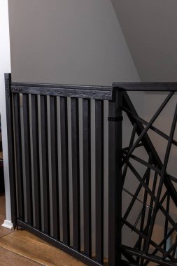 Dikey çıtçıtlı ve çapraz dizaynlı siyah metal kapı, tahta döşemeli ve nötr duvarlı bir merdivenin tepesine yerleştirilmiş..