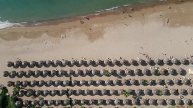 Hava manzarası, sıralanmış saman şemsiyeli ve güneşli yataklı bir plajı gösteriyor. Turkuaz dalgalar kumlu sahille buluşur, insanlar güneşin ve denizin tadını çıkarırlar..