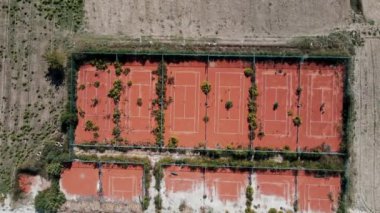 Yukarıdan birkaç kil tenis kortu görülüyor, bir ızgarada sıralanmış ve kırsal bir alanla çevrili yabani otlar ve küçük ağaçlarla kısmen gizlenmiş..