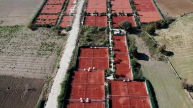Paralel sıralarda sıralanmış kırmızı kil tenis kortlarının hava perspektifi, tarlalar ve seyrek bitki örtüsüyle çevrili, oyuncular kibritlerle meşgul.
