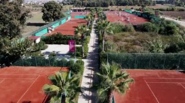 Palmiye ağaçlarıyla dolu bir dizi kil tenis kortu ve şemsiyeli masalar. Ağaçlarla ve açık alanlarla çevrili bir kortta bir kişi aktiftir..