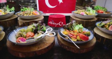 Etler, sebzeler ve 'Türkiye' ve 'Hilal Ay' ve yıldız sembollü kırmızı bir fes içeren ahşap platformlarda bir dizi Türk yemeği.