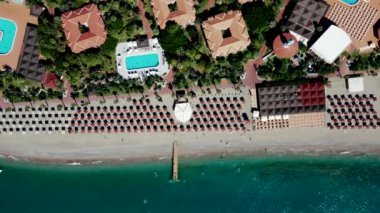 Canlı sahil beldesinde sıralı güneş pansiyonları, turkuaz deniz, küçük bir iskele, kırmızı kiremitli çatı binaları, yemyeşil bitki örtüsü ve çoklu yüzme havuzları yer alıyor..