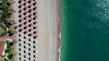Kırmızı çatılı şemsiyeler, palmiye ağaçları ve kırmızı çatılı bir yapıyla çevrili kumlu plajda sıralanır. Turkuaz deniz dalgaları kıyıya vuruyor, suda insanlar var..