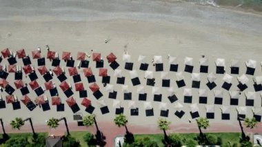 Alternatif renkli şemsiyeler, açık mavi yeşil su, küçük tekneler ve bir tatil köyü geçidi boyunca uzanan palmiye ağaçları olan bir sahili görüntüler..