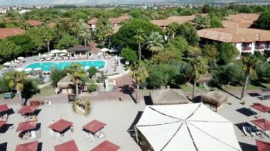 Bir tatil köyünde, tropik bitkilerle çevrili büyük bir havuz, şezlonglar ve şemsiyelerle çevrilidir. Çatılı yapılar ve kırmızı fayanslı binalar görülebilir..