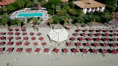 Sahile yakın bir tatil köyünde uzun bir iskele, kırmızı şemsiyeler ve güneş panzeleri bulunur. Kırmızı kiremitli çatı binaları ve saat kulesi yemyeşil ağaçlarla çevrili..