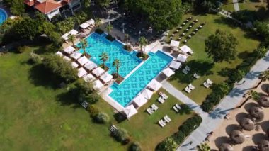 Bir tatil köyü havuzu, güneş şezlongları, şemsiyeler ve palmiye ağaçlarıyla çevrili geometrik bir tasarıma sahiptir. Yanı başında sazdan şemsiyeler olan kumlu bir sahil var..