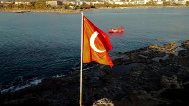 Önde büyük bir Türk bayrağı belirginleşirken, kayalık oluşumlar denize dökülüyor. Küçük bir tekne görünür, ve binalar uzak kıyılarda..