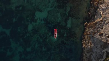 Kayalık bir sahil şeridinde, karanlık ve berrak sularda yüzen kırmızı bir tekne. Koyu mavi deniz, açık renkli kayalarla çelişiyor ve teknenin izolasyonunu vurguluyor..
