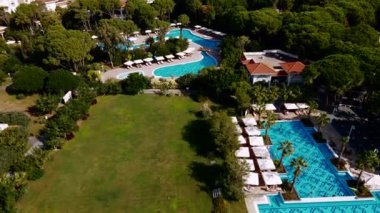 Büyük tatil beldesi, sarmal yüzme havuzları, güneş pansiyonları ve açık çimenli alanları olan bahçeler ve ağaçlarla çevrili kırmızı kiremitli çatı binaları içerir..