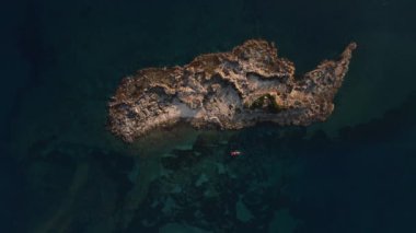 Havadan bakıldığında pürüzlü kenarları ve seyrek bitkileri olan küçük kayalık bir ada görülüyor. Adanın yakınındaki kırmızı bir kano koyu mavi suyla çelişiyor..