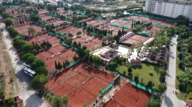 Ağaçlar ve ağlarla çevrili bir dizi kil tenis kortu kullanılıyor. Geniş bir bina ve yüksek binalar bir kentsel spor tesisini işaret ediyor.