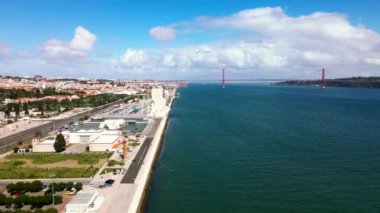 25 de Abril Köprüsü, Portekiz 'in Lizbon kentinde bulunan Tagus Nehri boyunca uzanır. Şehir manzarası parçalı bulutlu bir gökyüzü altında çeşitli mimari özelliklere sahiptir..